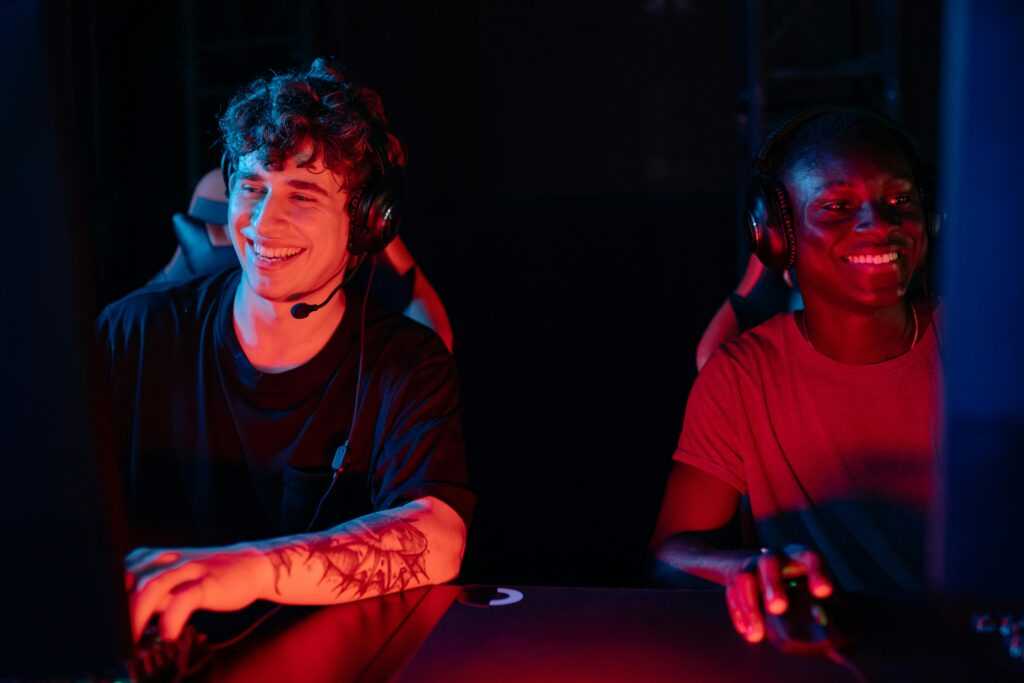 Smiling male gamers playing on PC with headsets in a dimly lit room.